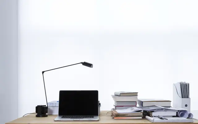 A laptop on a wooden desk beside a desk lamp and stacks of books and papers against a bright white wall