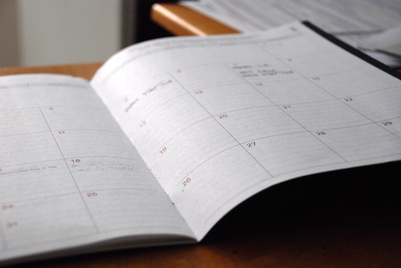 An open monthly planner on a wooden desk, its calendar grid showing dates with a few handwritten notes